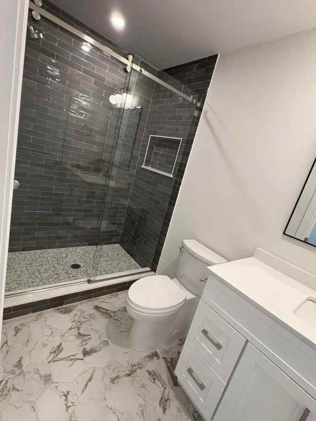 A walk-in shower with dark subway tile and a glass sliding door, beside a white vanity