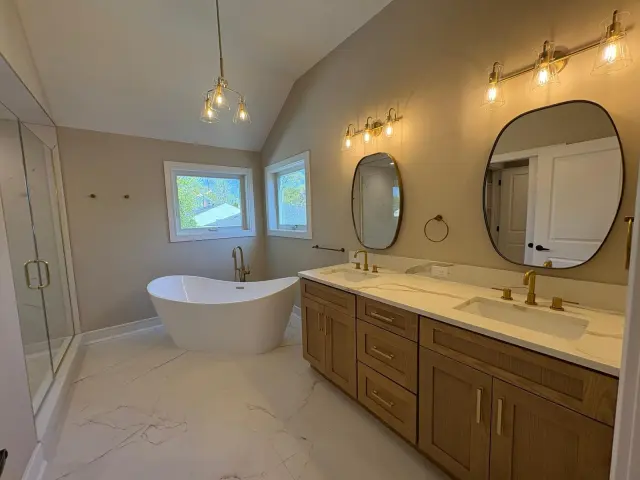 A double vanity with oval mirrors and brass lights, and a freestanding tub under the window