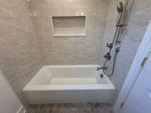 A white tub in gray marble-look tile, with a recessed niche and hand shower