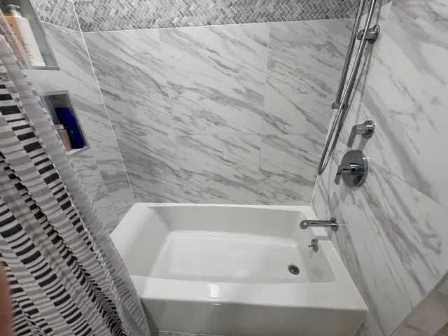 A white tub in a marble-tiled bath, with a hand shower and recessed niches